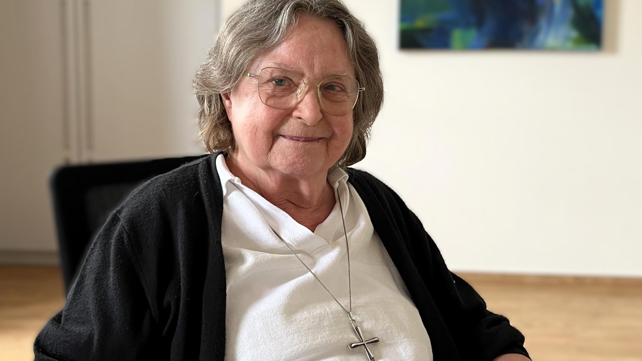 Ältere Frau mit grauen Haaren, Brille und einem großen silbernen Kreuz um den Hals sitzt an einem Tisch und lächelt in die Kamera