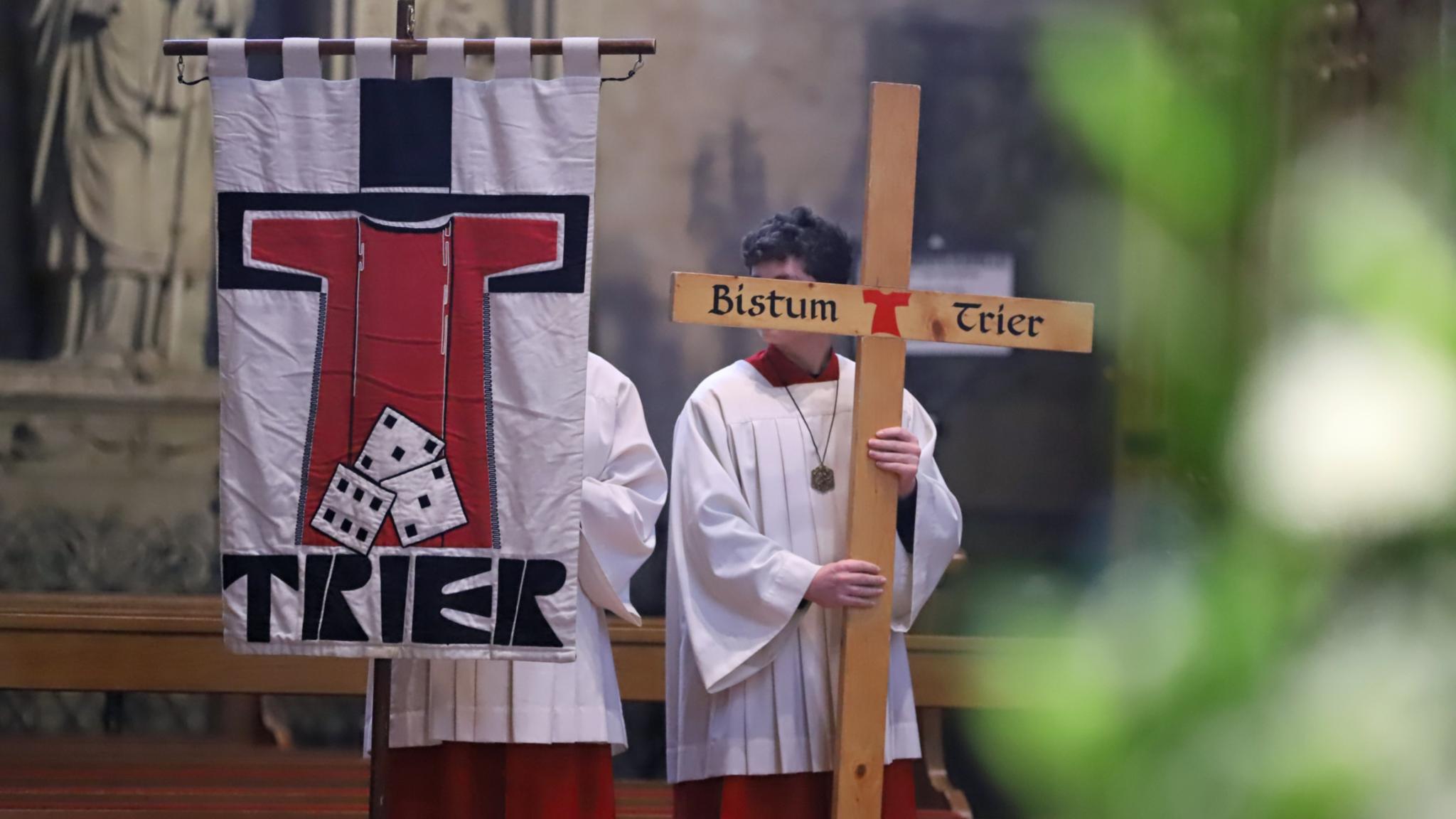Zwei Messdiener in einer Messe; einer hält die Pilgerfahne mit der Aufschrift 'Trier' hoch, der andere ein Kreuz, auf dem Bistum Trier steht.