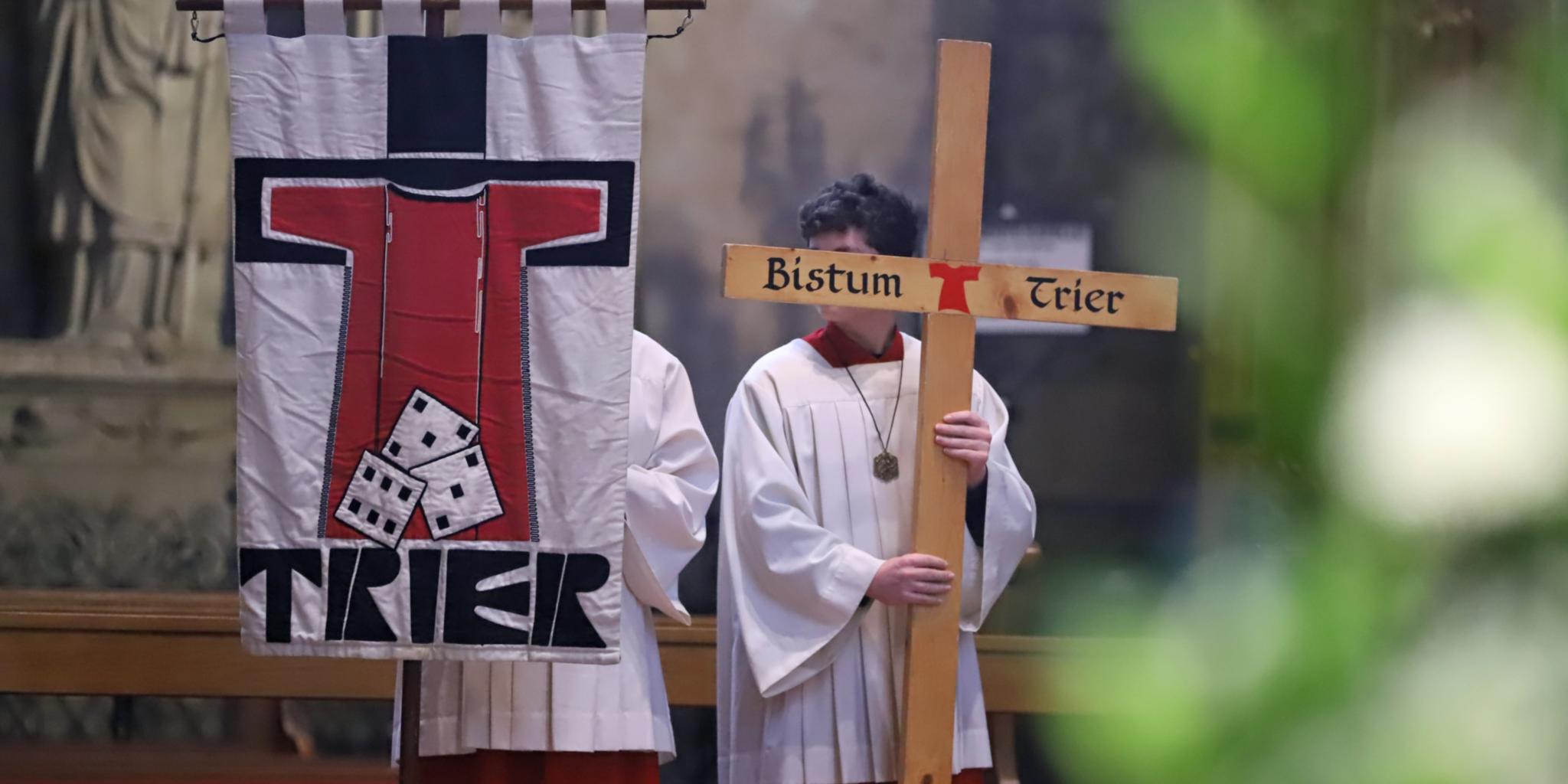 Zwei Messdiener in einer Messe; einer hält die Pilgerfahne mit der Aufschrift 'Trier' hoch, der andere ein Kreuz, auf dem Bistum Trier steht.
