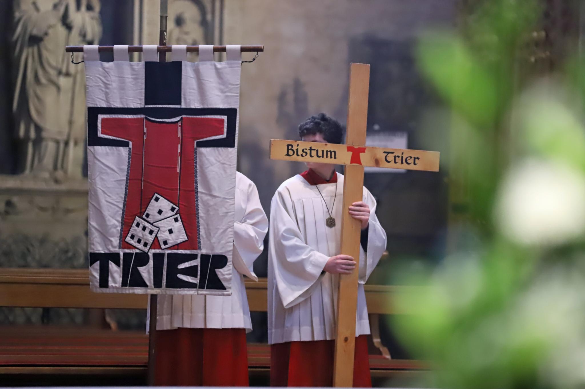 Zwei Messdiener in einer Messe; einer hält die Pilgerfahne mit der Aufschrift 'Trier' hoch, der andere ein Kreuz, auf dem Bistum Trier steht.