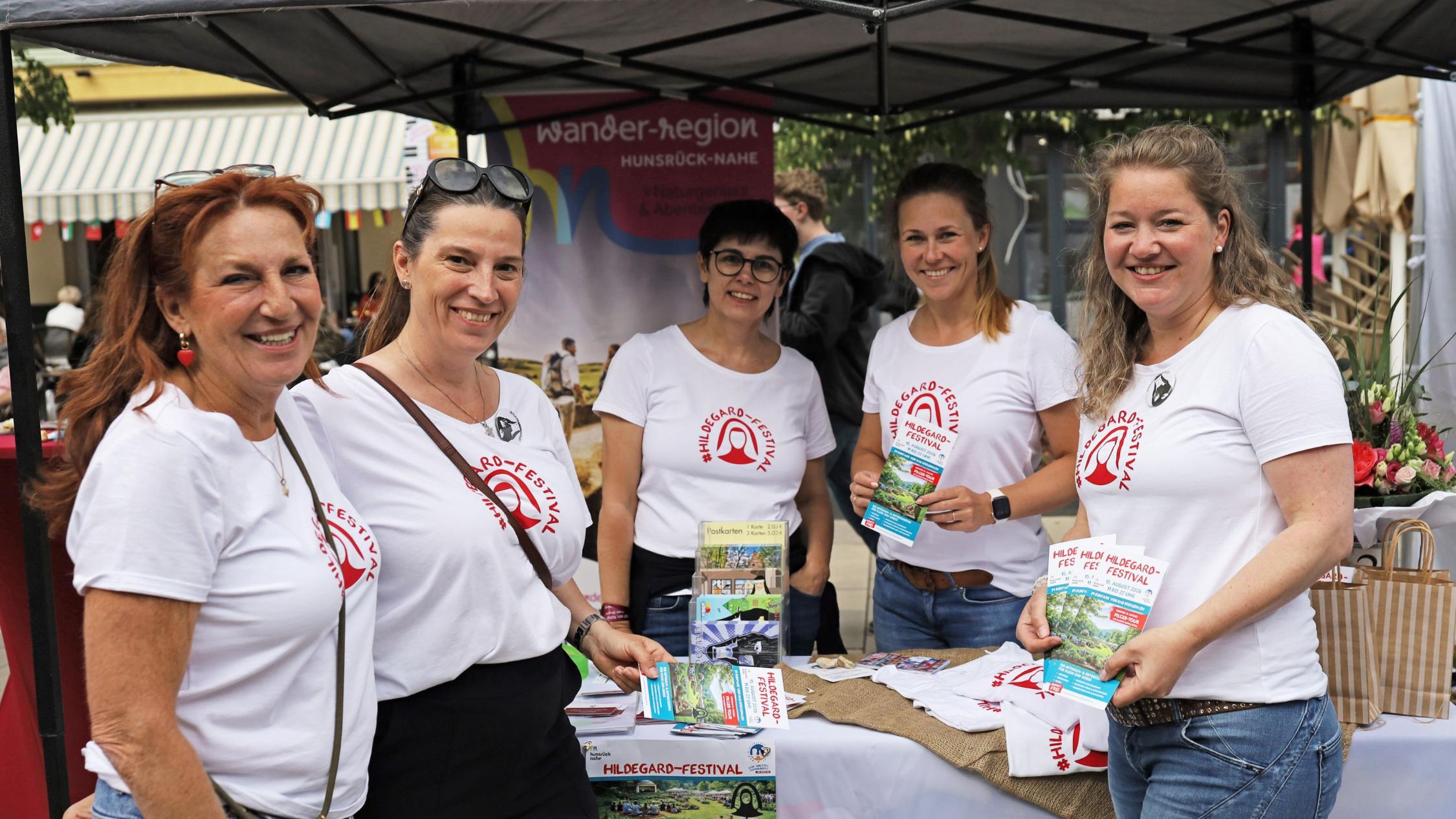 5 Frauen mit T-Shirts (Aufschrift #Hildegard-Festival) stehen am Infostand
